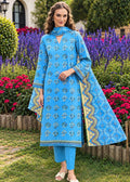 A woman stands outdoors in a garden wearing a bright blue three-piece traditional outfit with matching pants and a draped dupatta. The outfit features intricate floral prints and gold-colored border details.
