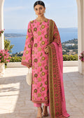 A woman stands in a sunlit outdoor terrace wearing a bright pink traditional outfit with ornate gold and green floral embroidery and a matching embroidered dupatta. The background shows a coastal sea view through white arches.