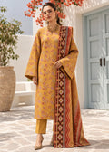 A woman models a traditional Indian salwar kameez suit in mustard yellow with intricate floral embroidery, paired with matching yellow trousers and a red and gold patterned dupatta, set against a Mediterranean-style courtyard background.