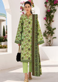 A woman models a light green three-piece traditional outfit with intricate black and gold floral patterns, paired with matching green pants and a patterned dupatta, set against a white architectural background with climbing pink flowers.