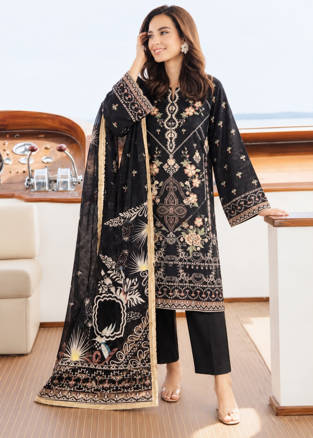 A woman models a black three-piece traditional outfit with intricate gold and beige embroidery, featuring a long kurta, black pants, and a matching dupatta, set against a luxurious yacht cabin background.
