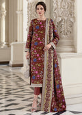 A woman models a traditional Indian salwar kameez suit in a grand indoor setting with classical columns. The outfit consists of a brown long kurta decorated with colorful floral prints and a wide embroidered border, paired with matching brown trousers and a silver clutch.
