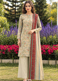 A woman stands outdoors in a garden wearing a traditional three-piece salwar kameez suit. The set features a beige kurta with a light blue and red floral print, matching beige wide-leg trousers, and a red dupatta with blue and green borders.