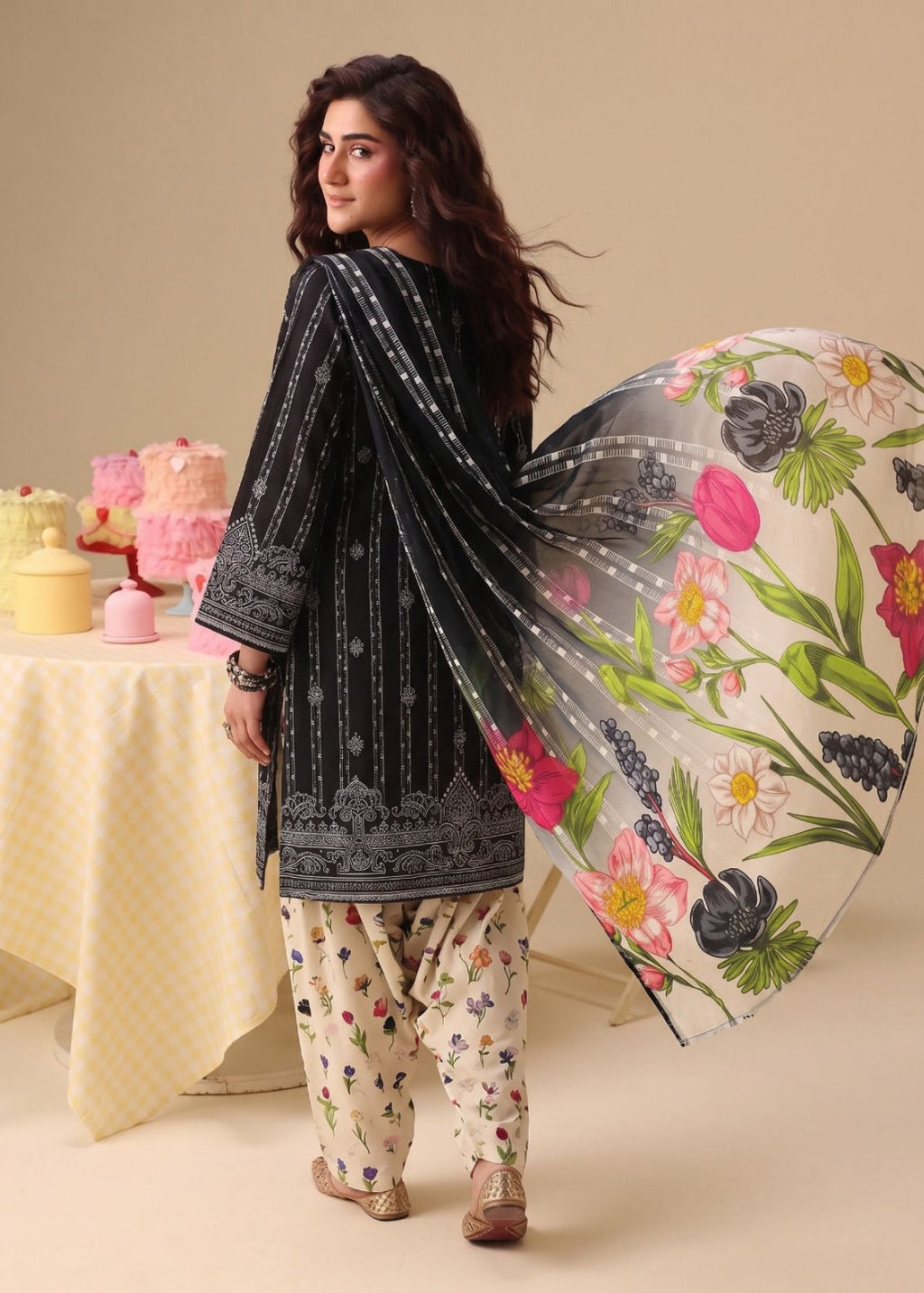 A woman modeling a traditional South Asian outfit, featuring a black embroidered kurta, cream floral pants, and a sheer black dupatta with a vibrant multicolor floral print, set against a warm beige background with a decorated table in the background.