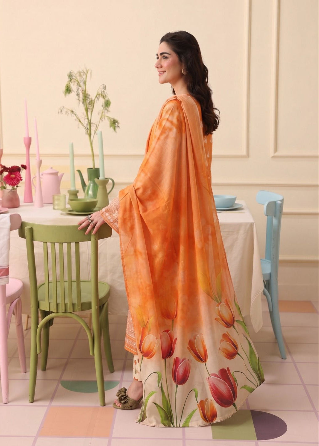 A woman models a peach and orange printed dupatta and matching skirt set with a tulip floral design, standing in a pastel-colored dining room setting.