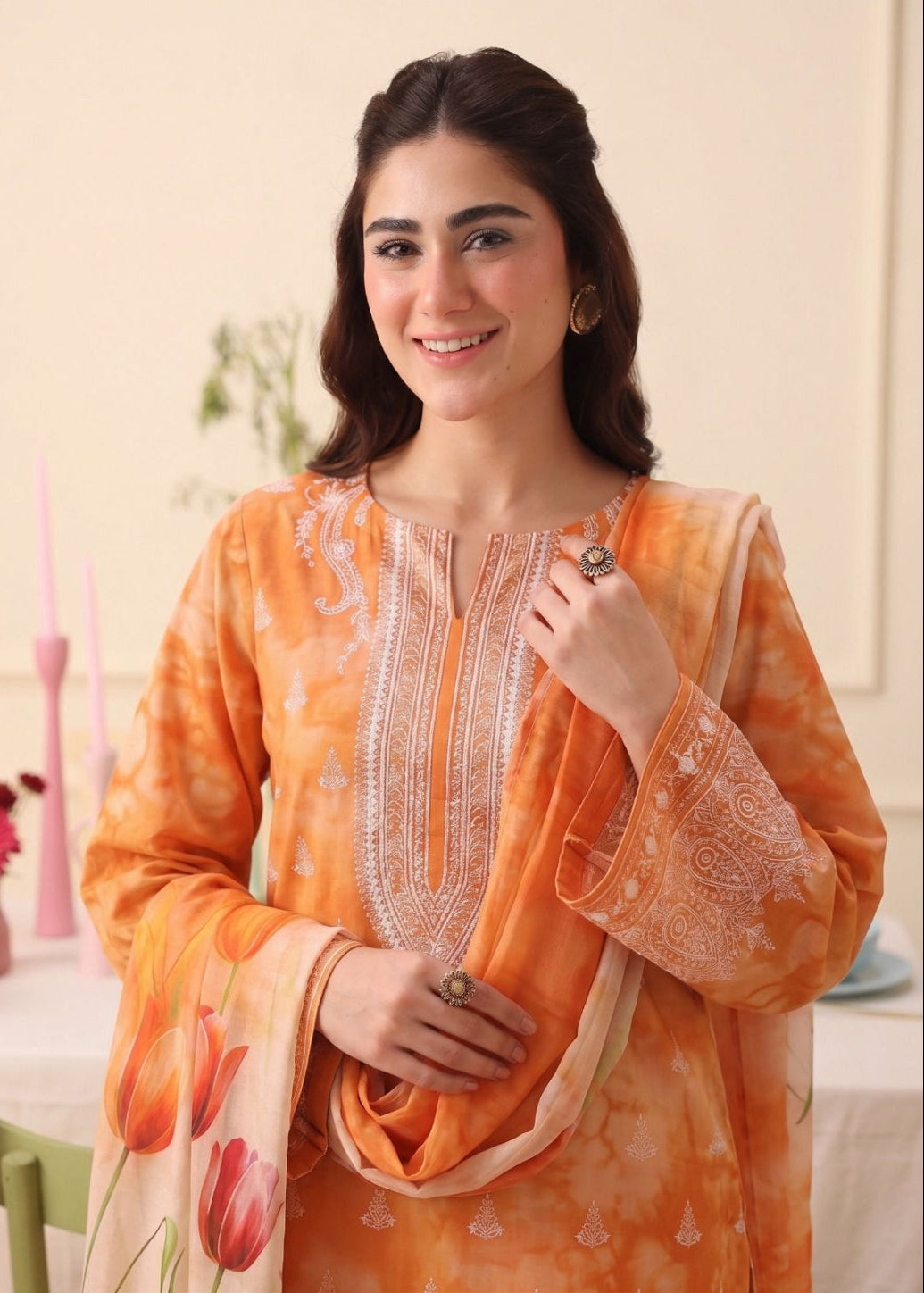 A woman modeling a traditional orange salwar kameez suit with white embroidery and a matching printed dupatta featuring orange and pink tulip motifs, set against a soft beige indoor background.