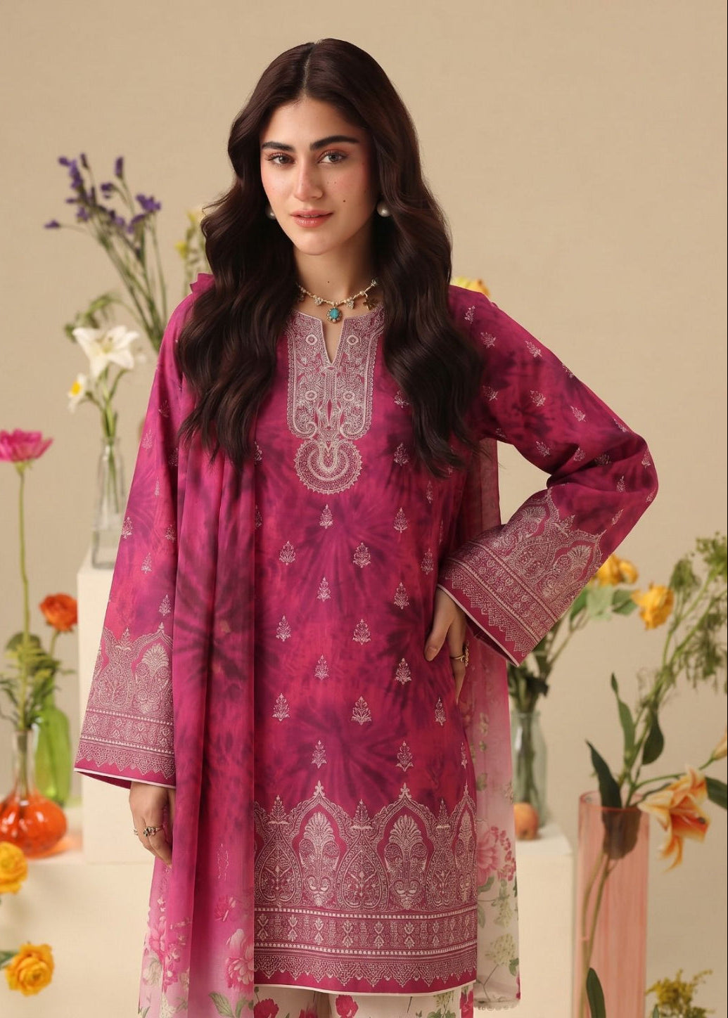 A woman modeling a magenta pink traditional South Asian salwar kameez suit with intricate silver mirror work embroidery on the neckline, sleeves, and hem, paired with a matching dupatta. The background is a warm beige studio setting decorated with floral arrangements.