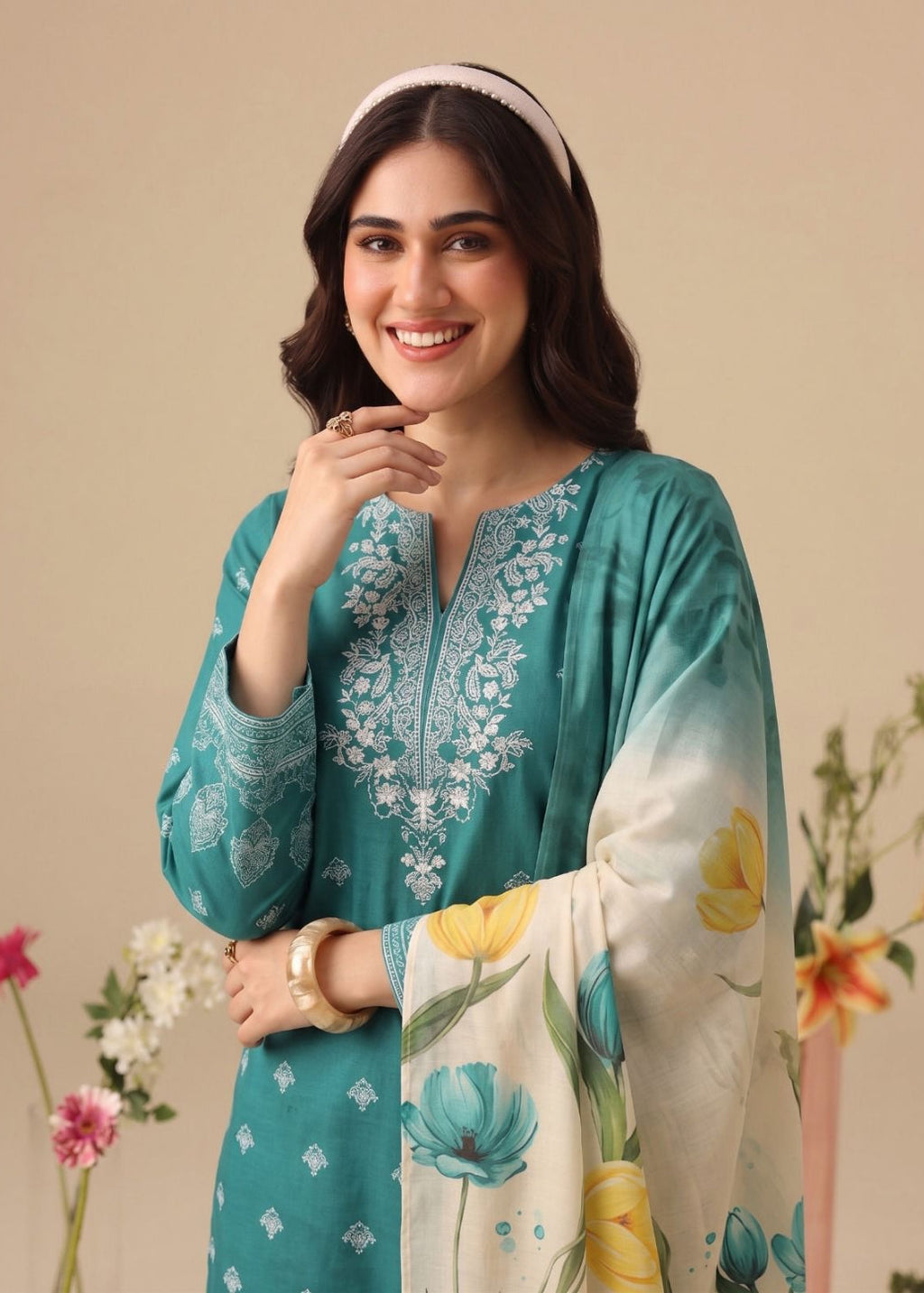 A woman modeling a teal green ethnic kurta with white embroidery, paired with a cream dupatta featuring a blue and yellow floral print, set against a warm beige background with decorative flowers.