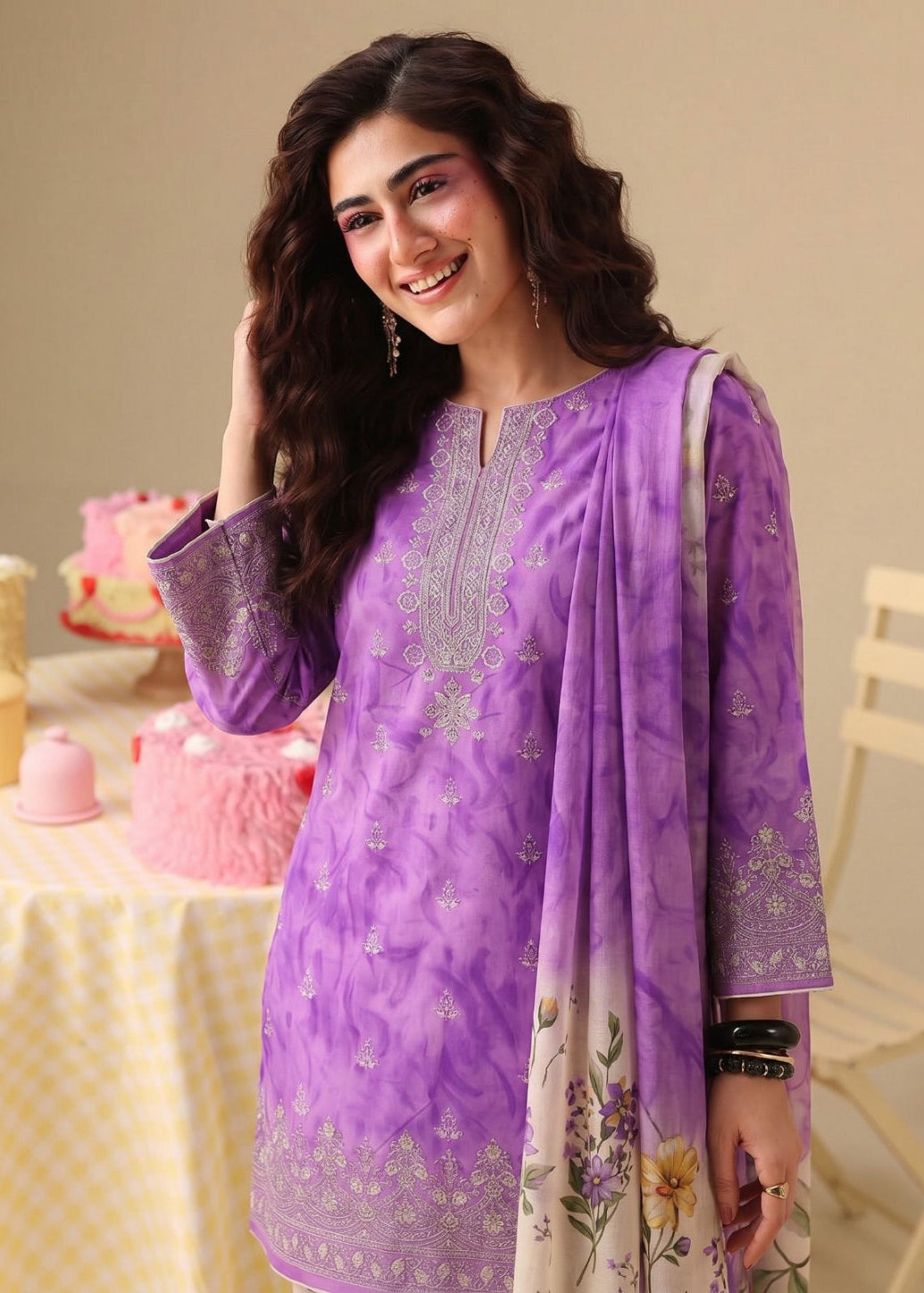 A woman modeling a purple three-piece salwar kameez suit with silver embroidery and a floral printed dupatta, set against a warm indoor background with a decorated table.