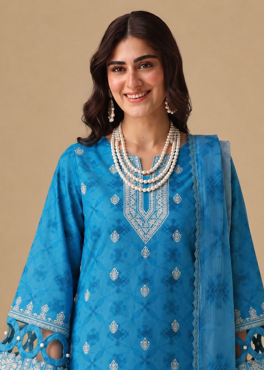A woman modeling a bright blue traditional outfit with intricate silver embroidery, featuring a matching sheer blue dupatta, set against a solid beige background.