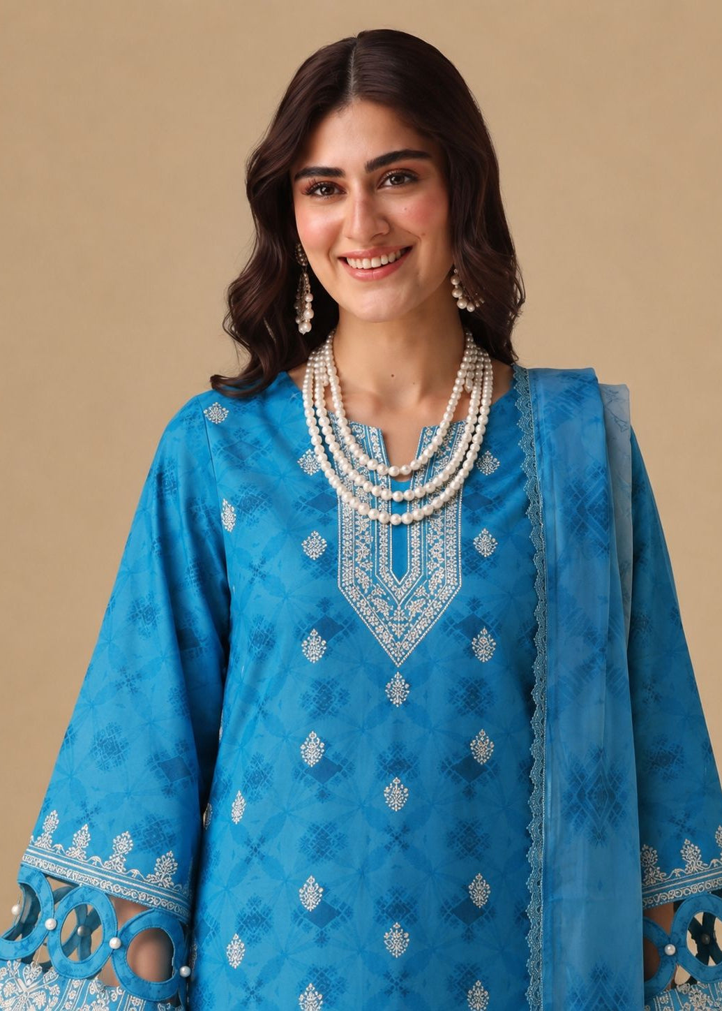 A woman modeling a bright blue traditional outfit with intricate silver embroidery, featuring a matching sheer blue dupatta, set against a solid beige background.