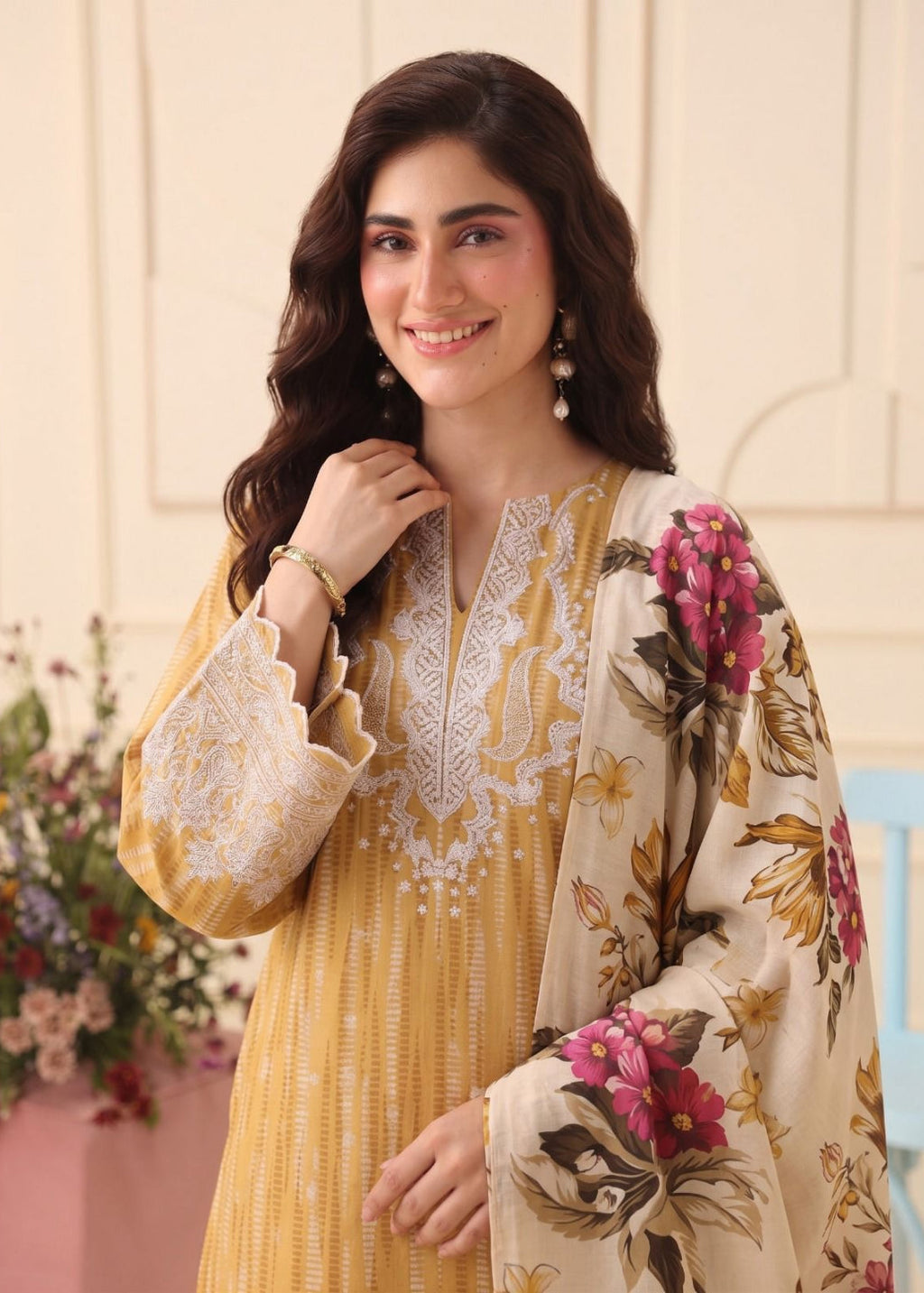 A woman modeling a traditional South Asian outfit featuring a mustard yellow embroidered kurta with white lace detailing, paired with a beige dupatta decorated with pink and brown floral patterns.