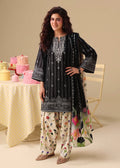 A woman models a black embroidered kurta with a floral print dupatta, standing in front of a beige background with a decorated table and chair.
