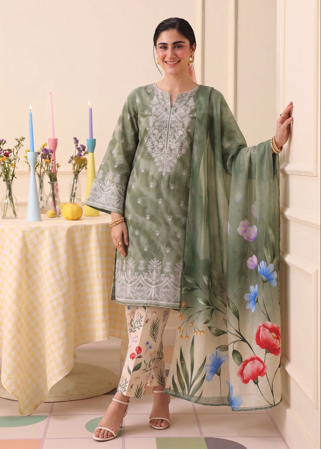 A woman models a three-piece traditional South Asian salwar kameez set. The set includes a sage green kurta with white embroidery, matching printed trousers, and a sheer sage green dupatta decorated with colorful floral prints.