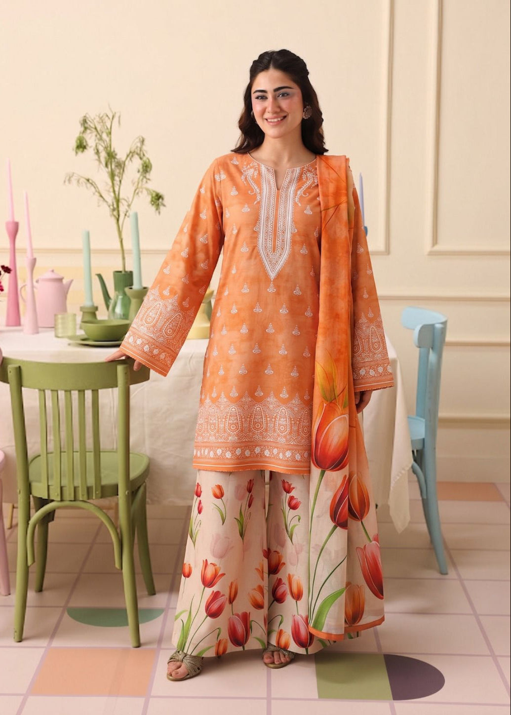 A woman modeling a traditional three-piece salwar kameez suit in a pastel-colored room. The set features an orange kurta with white embroidery, matching wide-leg trousers with a tulip print, and a coordinating orange dupatta.