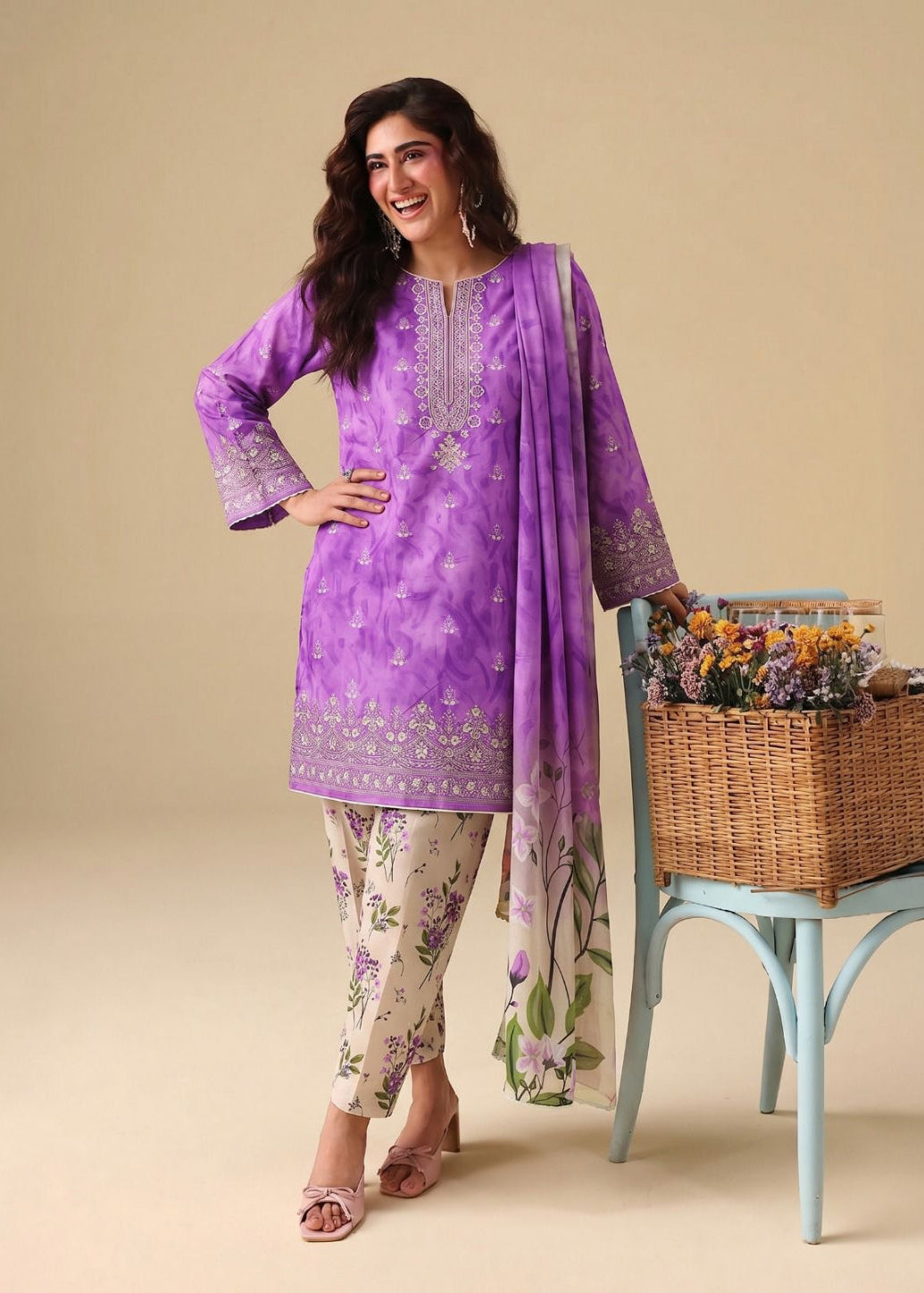 A woman models a traditional three-piece salwar kameez suit in a studio setting. The set features a lavender purple printed and embroidered kurta, beige floral printed pants, and a matching purple dupatta draped over her shoulder.