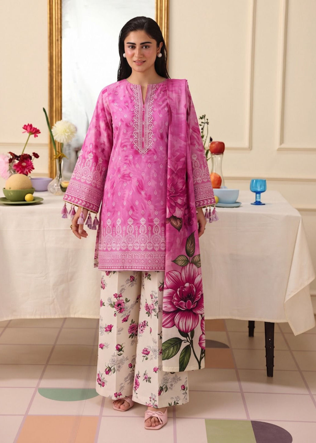 A woman modeling a pink traditional three-piece salwar kameez suit with floral prints, standing in a decorated room with a table and mirror in the background.