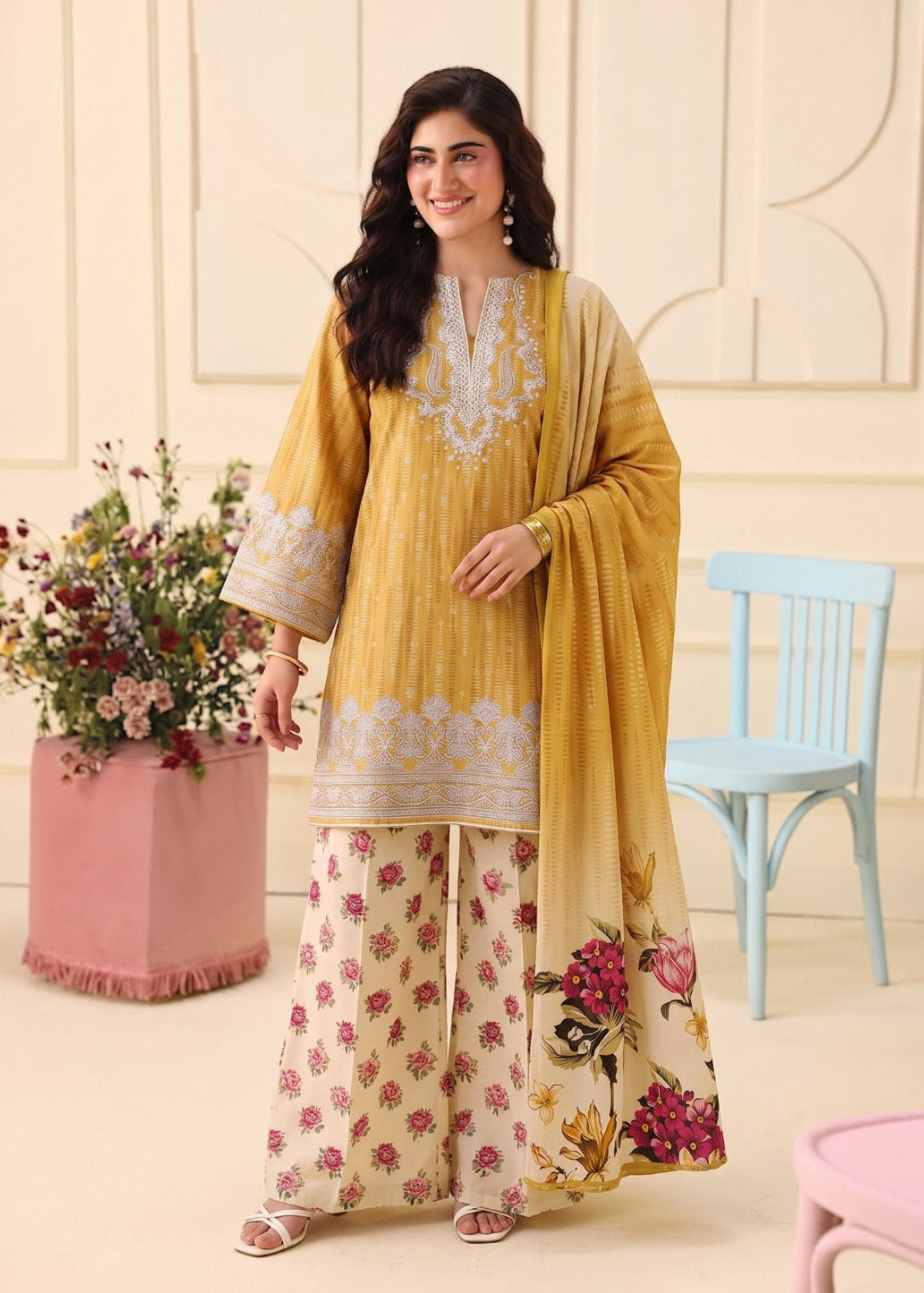 A woman models a traditional three-piece salwar kameez suit. The outfit consists of a mustard yellow embroidered tunic, wide-leg beige trousers with pink floral prints, and a matching yellow-to-beige gradient dupatta with floral embroidery and a butterfly print.