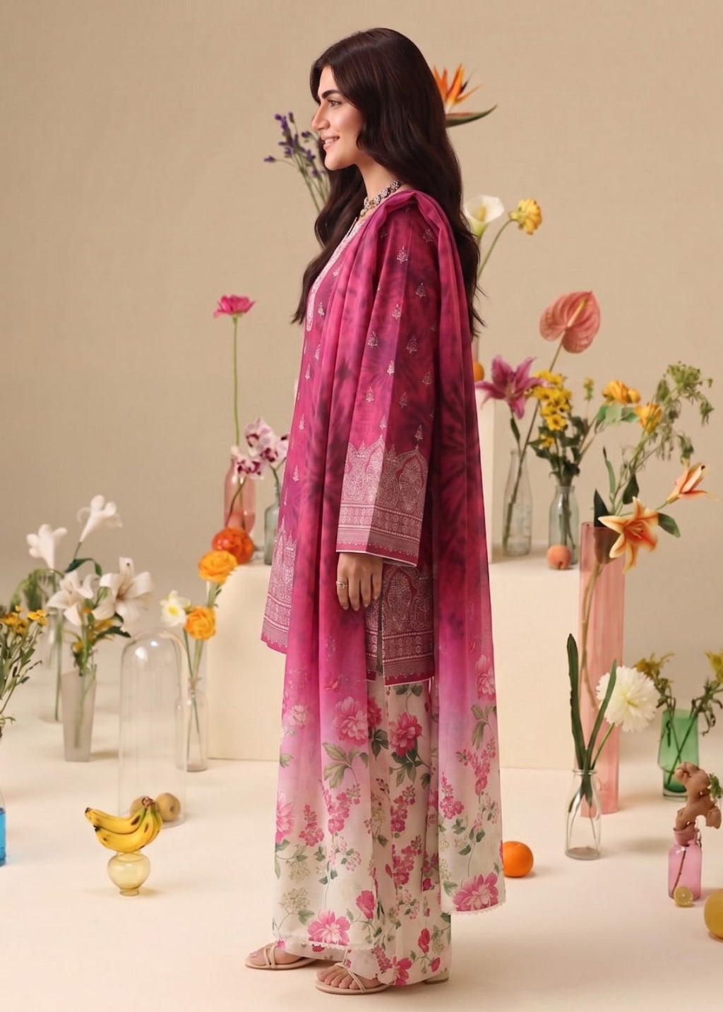 A woman modeling a three-piece pink and red floral printed salwar kameez suit with a matching dupatta, standing in a studio decorated with colorful flower vases.