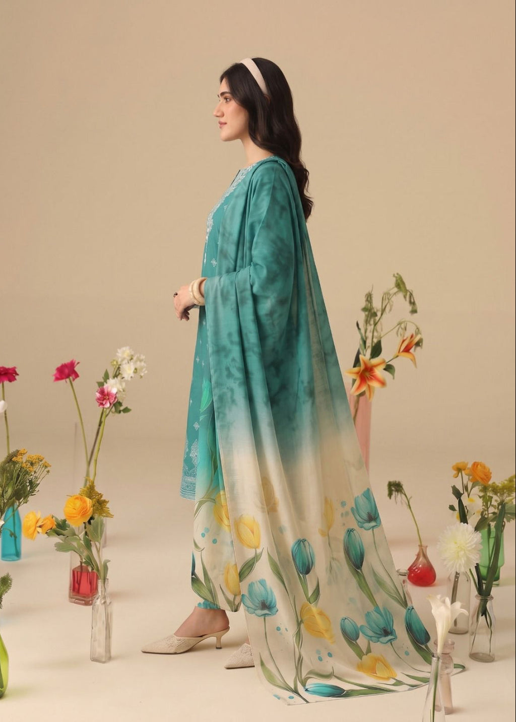 A woman models a traditional South Asian salwar suit set featuring a teal and white gradient tunic with a matching printed dupatta. The set is styled with floral arrangements in glass vases against a solid beige studio backdrop.