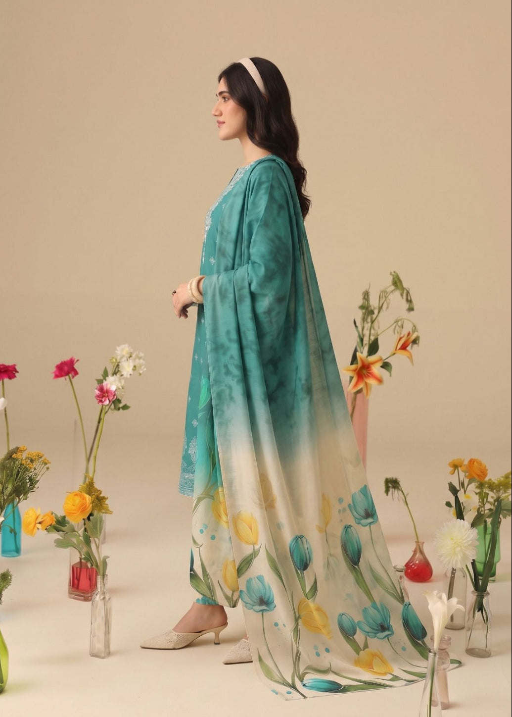 A woman models a traditional South Asian salwar suit set featuring a teal and white gradient tunic with a matching printed dupatta. The set is styled with floral arrangements in glass vases against a solid beige studio backdrop.