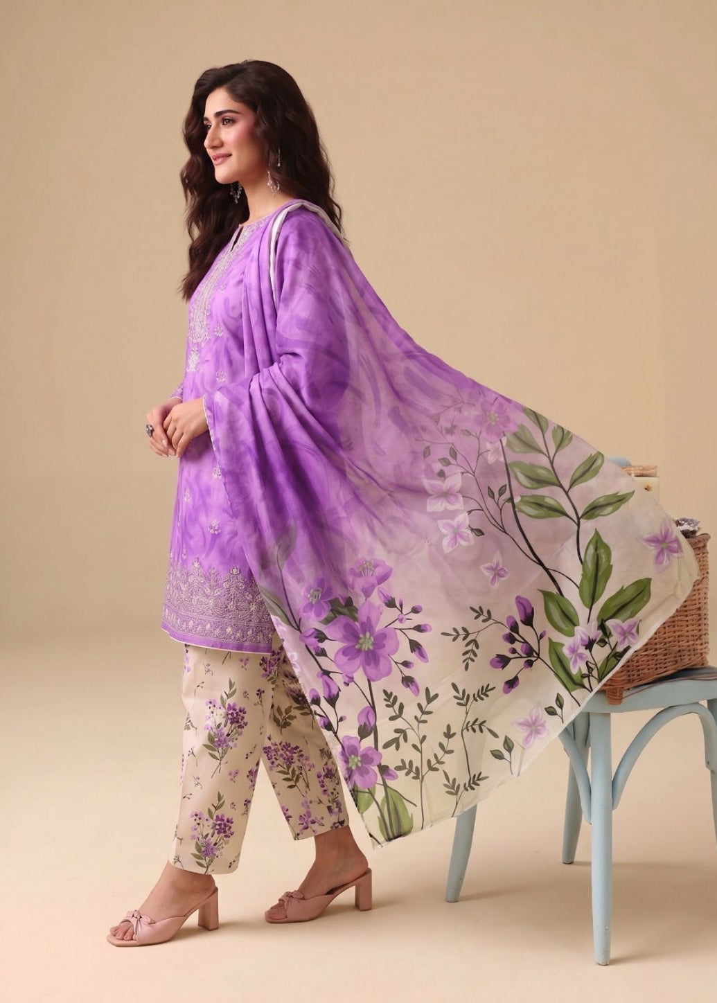 A woman models a traditional three-piece salwar kameez suit in lavender and beige with floral prints, featuring a matching sheer purple dupatta with green leafy designs, posed next to a light blue stool.