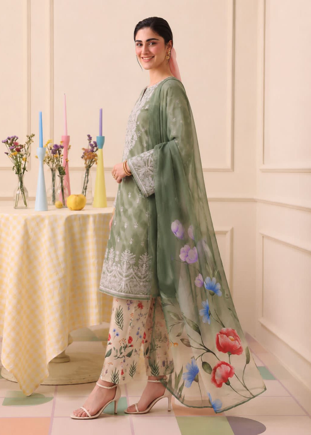 A woman models a traditional three-piece salwar kameez set in a decorated indoor setting. The set features a sage green embroidered kameez top with a matching sheer floral dupatta, paired with beige floral printed trousers.
