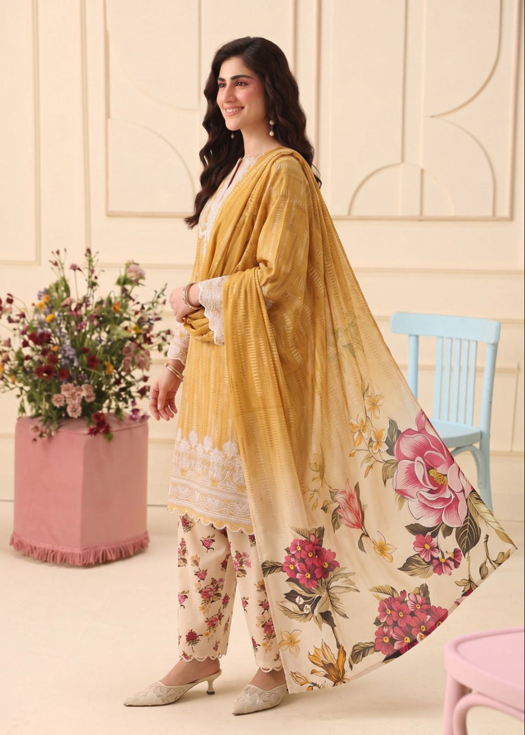 A woman models a traditional South Asian salwar kameez suit set. The outfit features a mustard yellow tunic with white floral embroidery, beige floral pants, and a matching yellow dupatta with a large pink and red floral print.