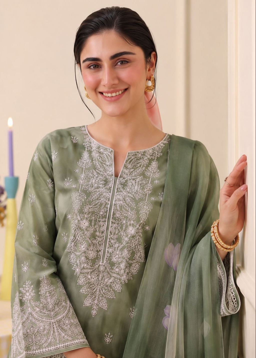A woman wearing a sage green embroidered traditional outfit with a matching sheer dupatta, standing indoors against a light beige wall.