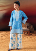 A woman modeling a light blue embroidered kurta with matching wide-leg pants featuring a blue floral print on a cream background, set against a warm desert-like backdrop with a wooden chair.