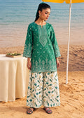 A woman stands on a sandy beach wearing a green embroidered kurta with matching wide-leg printed pants. The background shows the ocean, a table with a cream cloth, and part of a yellow umbrella.
