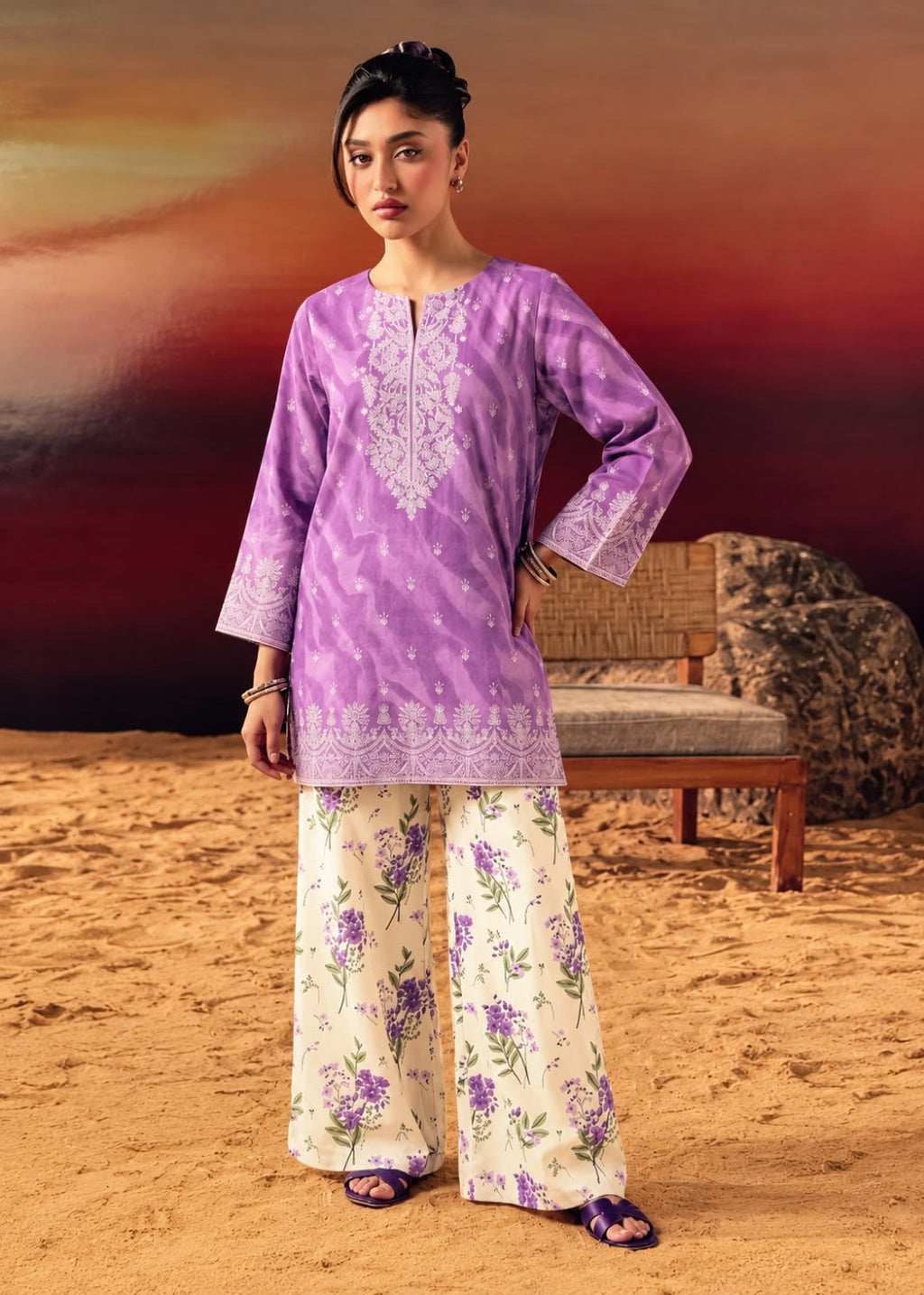 A woman modeling a lavender purple kurta with white floral embroidery and matching wide-leg pants featuring a purple floral print on a cream background, set against a desert landscape backdrop with a wooden bench.