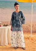 A woman stands on a sandy beach wearing a dark blue printed kurta with white embroidery and wide-leg cream floral pants. A table with a cream draped cloth, a yellow umbrella, and the ocean are visible in the background.