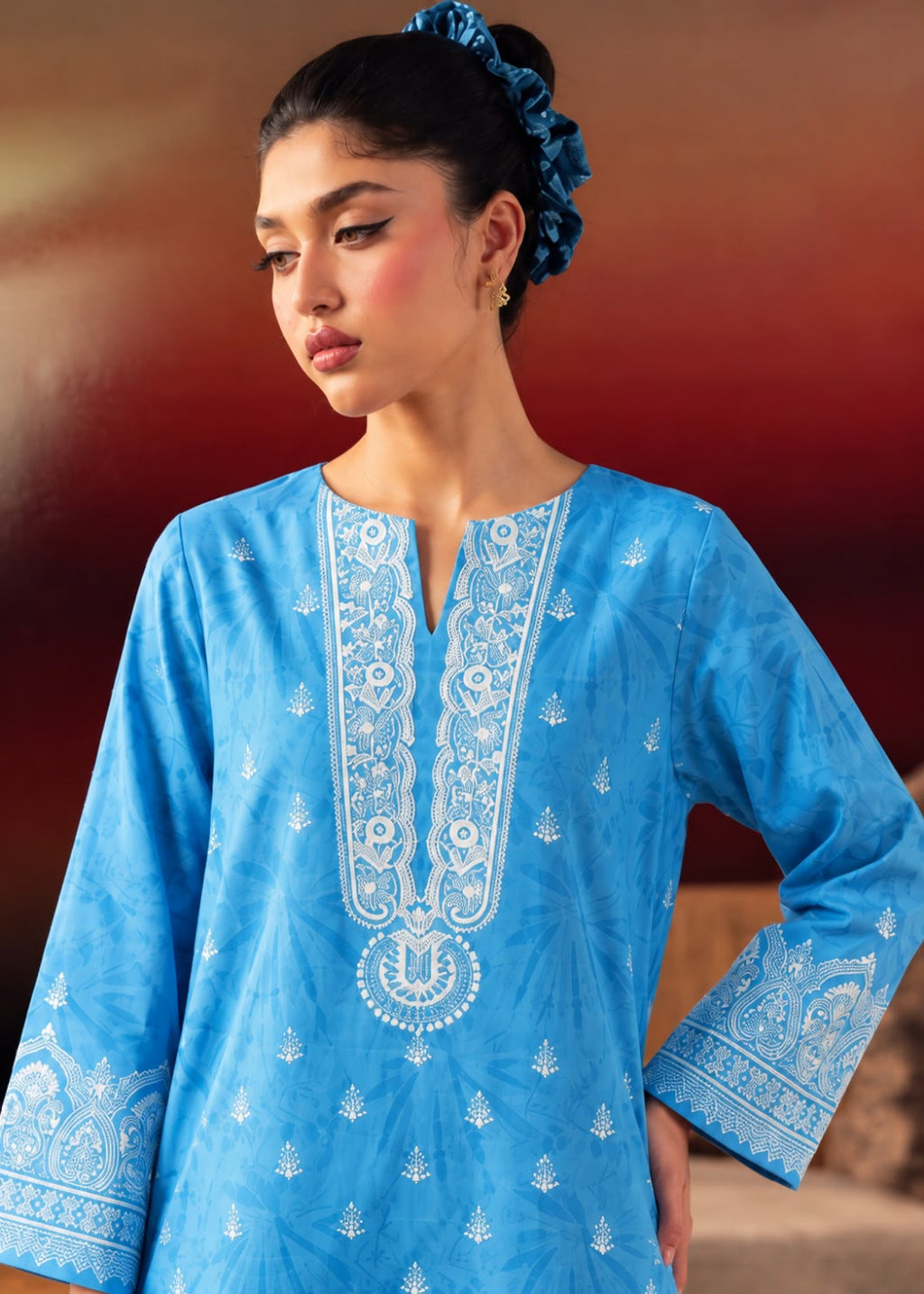 A woman wearing a bright blue traditional tunic with intricate white embroidery around the neckline and cuffs, posed against a warm brown and red gradient background.