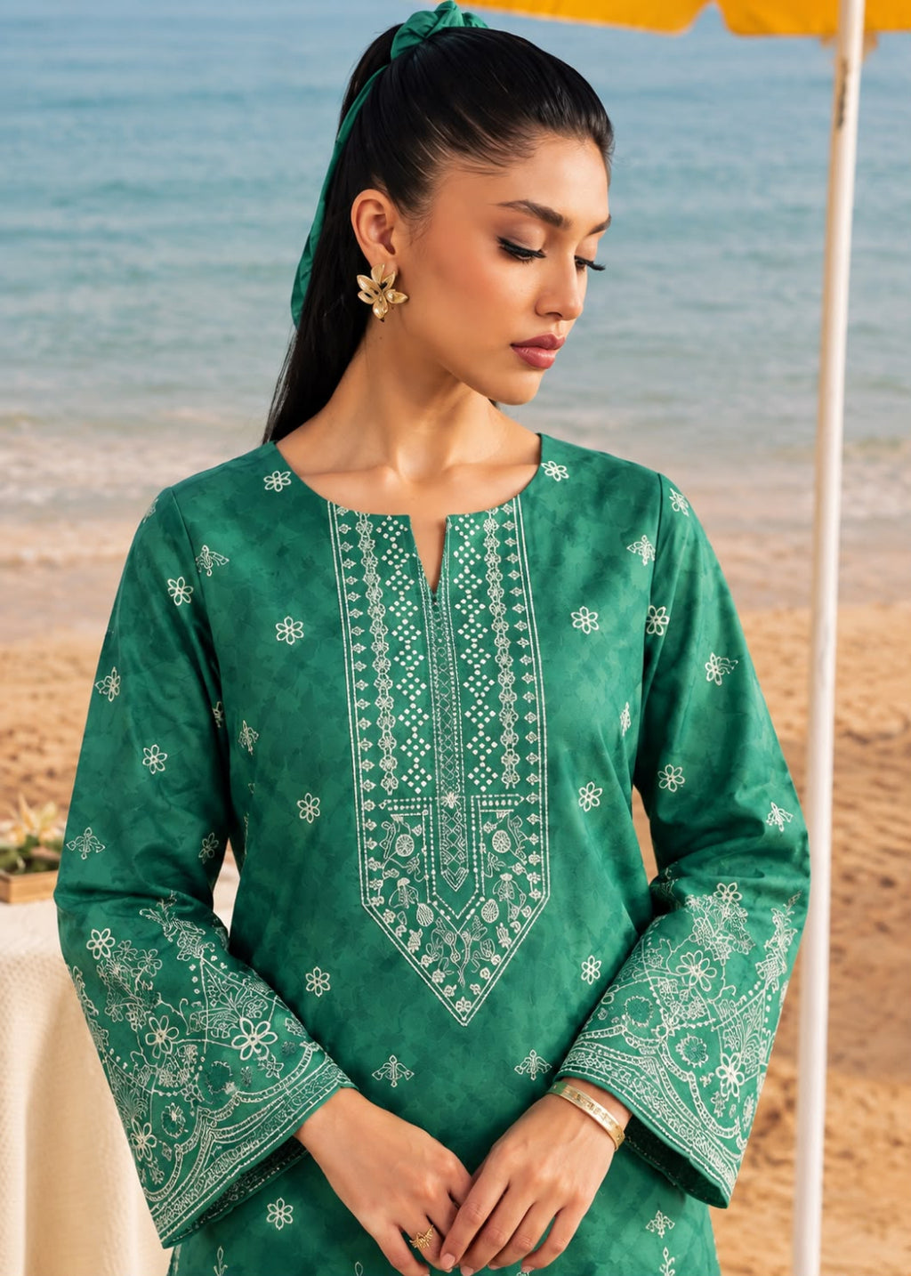 A woman wearing a green embroidered kurta stands outdoors near a beach, with ocean water and sand visible in the background.