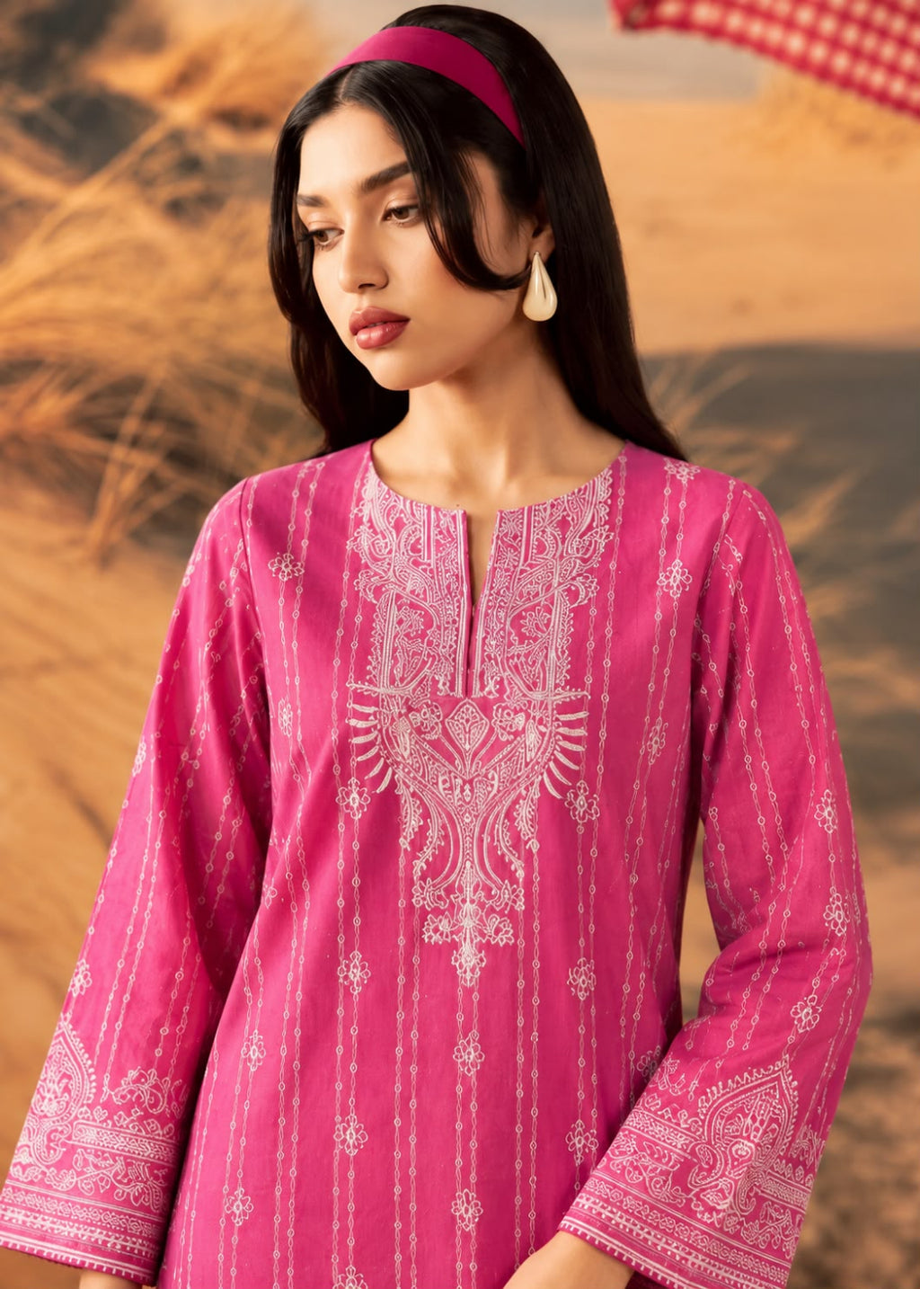 A woman wearing a bright pink traditional tunic with white embroidered patterns, set against a blurred desert background.