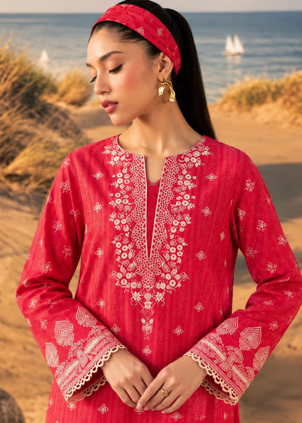 A woman wearing a red embroidered long kurta with a matching red headband, standing on a sandy beach with the ocean in the background.