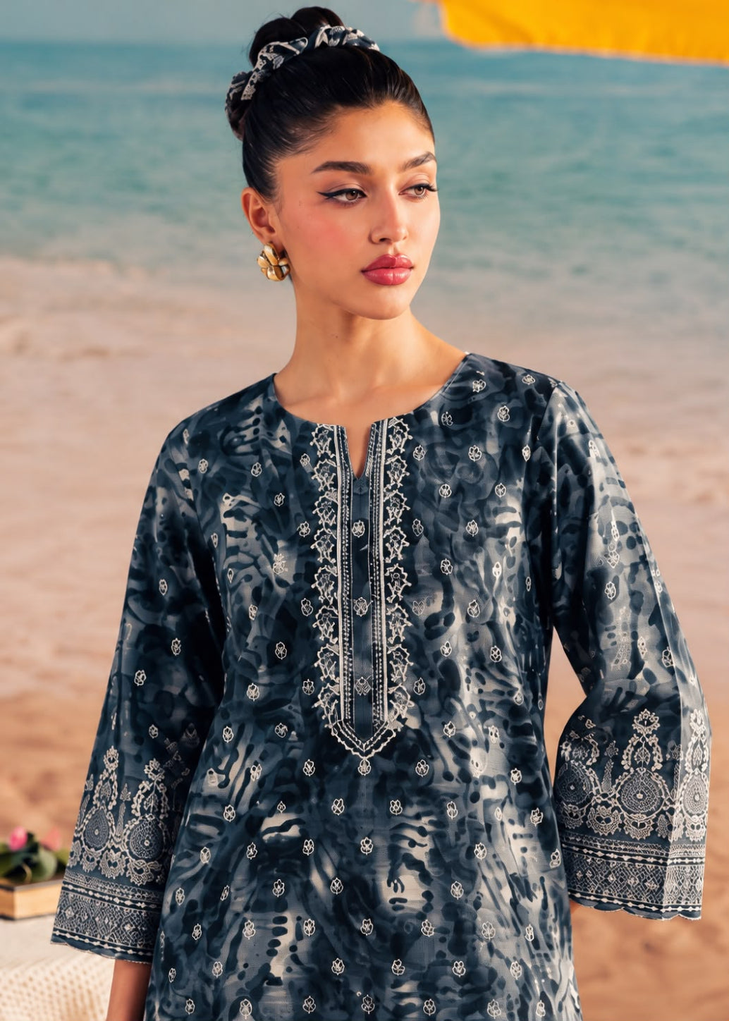 A woman modeling a dark blue-gray patterned tunic with white embroidery on a beach background.