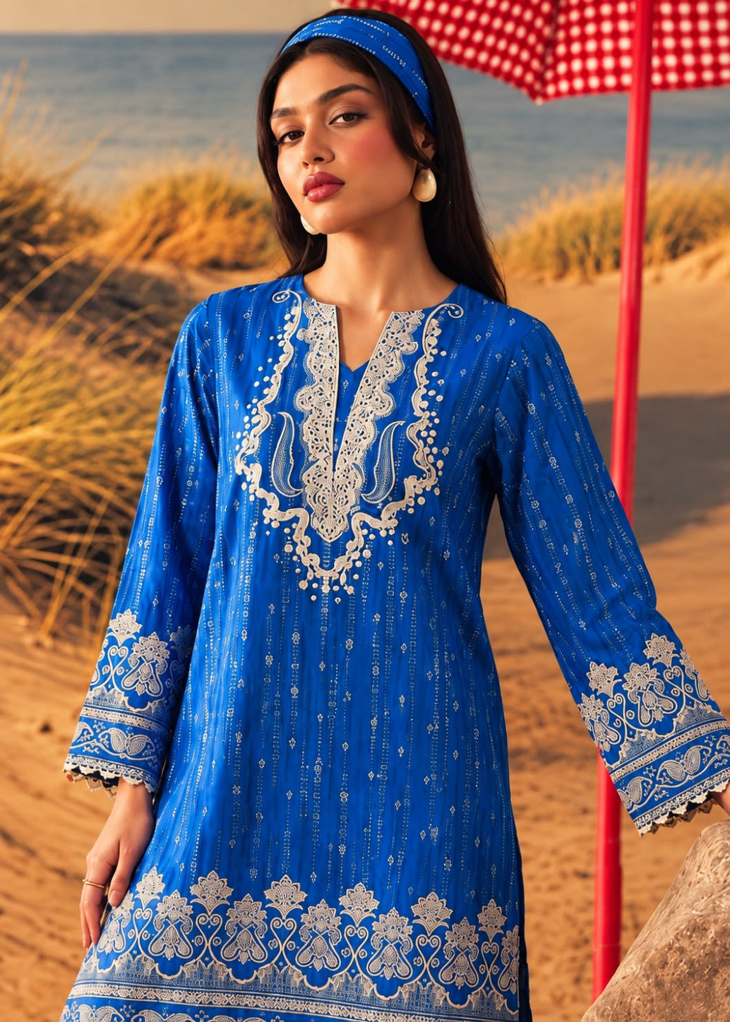 A woman modeling a royal blue embroidered kurta with white floral patterns, a matching blue headband, and a red-and-white gingham umbrella in the background on a sandy beach.