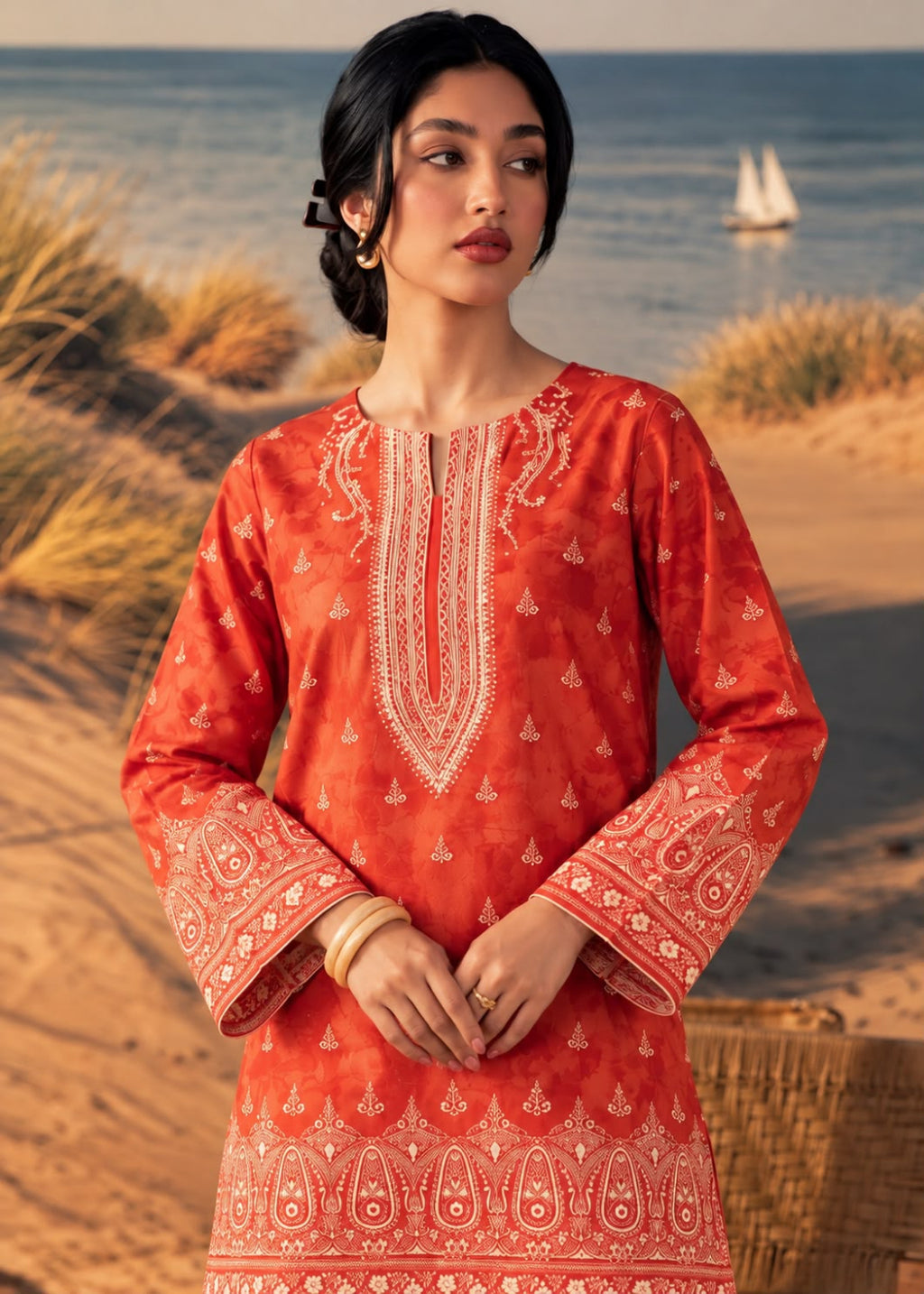 A woman wearing a coral-orange embroidered kurta stands on a sandy beach with the ocean and dunes in the background. The kurta features intricate white and light pink embroidery concentrated on the neckline, sleeves, and hem.