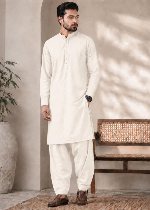 A man modeling a traditional white kurta and pant set in a warm, sunlit indoor setting with a textured wall, potted plant, and wooden bench.