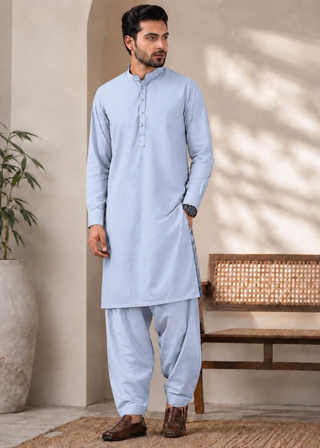 A man wearing a light blue traditional shirtdress (kurta and pant set) standing in a styled indoor setting with a bench and potted plant.