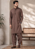 A man wearing a brown long-sleeve kurta and matching loose trousers, standing in a styled indoor setting with a bench, potted plant, and textured wall.
