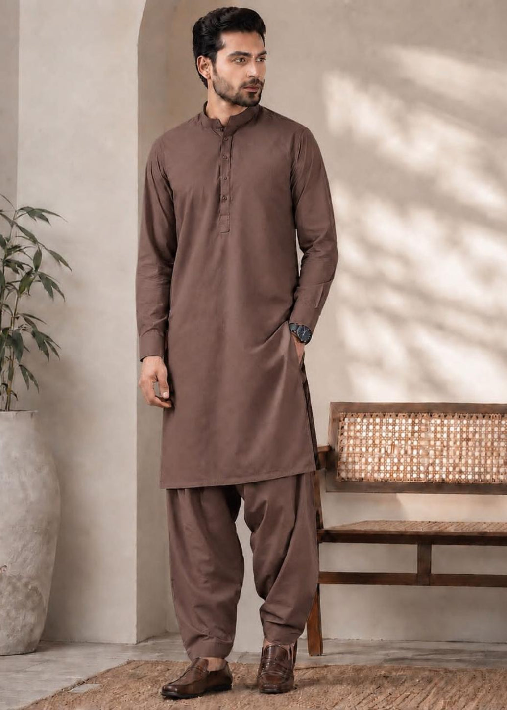 A man wearing a brown long-sleeve kurta and matching loose trousers, standing in a styled indoor setting with a bench, potted plant, and textured wall.