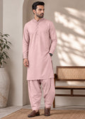 A man wearing a light pink traditional kurta and pant set, standing in a styled indoor setting with a wooden bench, potted plant, and textured rug.