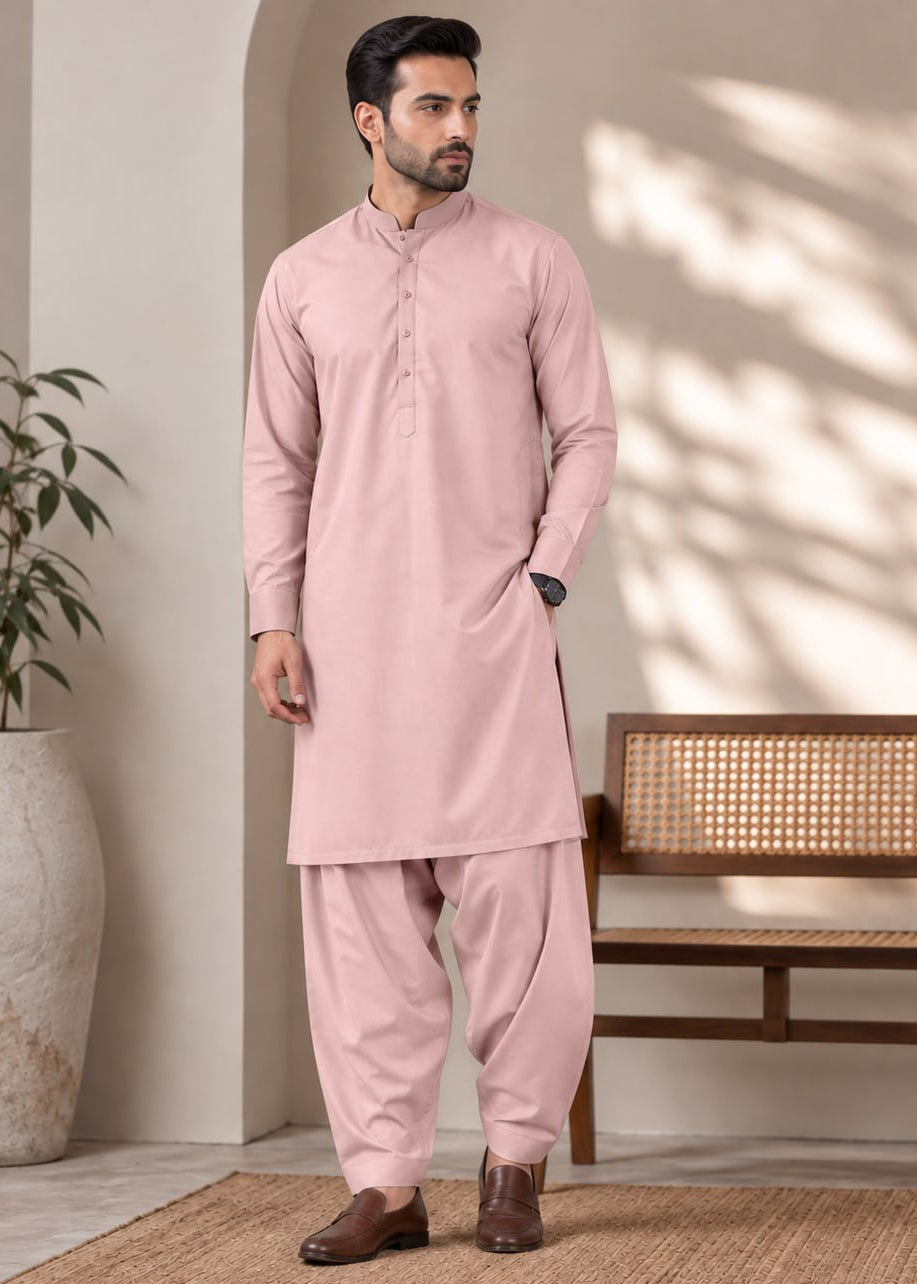 A man wearing a light pink traditional kurta and pant set, standing in a styled indoor setting with a wooden bench, potted plant, and textured rug.