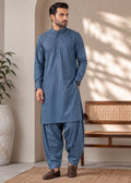 A man wearing a slate blue traditional kurta and pant set, standing in a styled indoor setting with a wooden bench, a potted plant, and a woven rug.