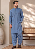 A man wearing a light blue traditional kurta and pant set stands in a styled indoor setting with a bench, potted plant, and textured rug.