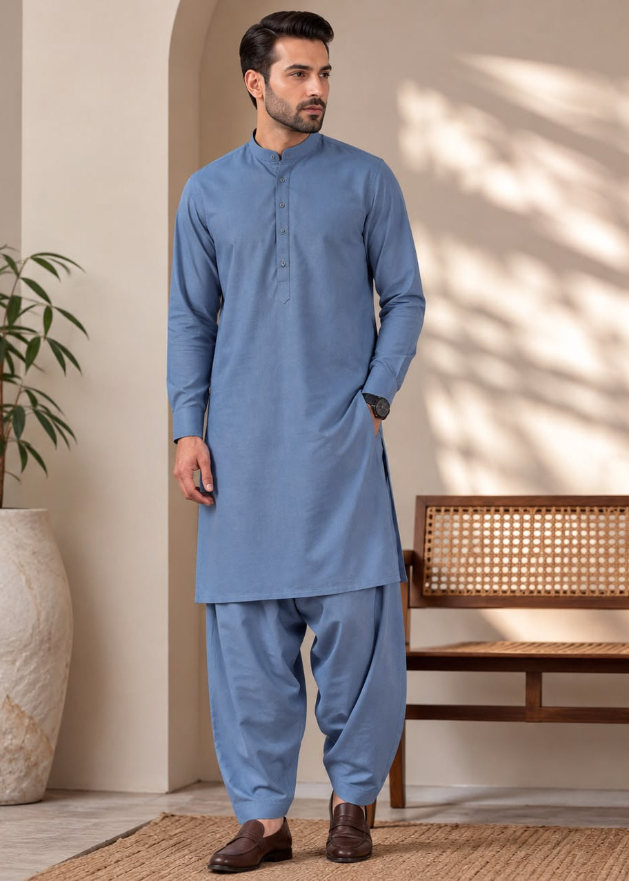 A man wearing a light blue traditional kurta and pant set stands in a styled indoor setting with a bench, potted plant, and textured rug.