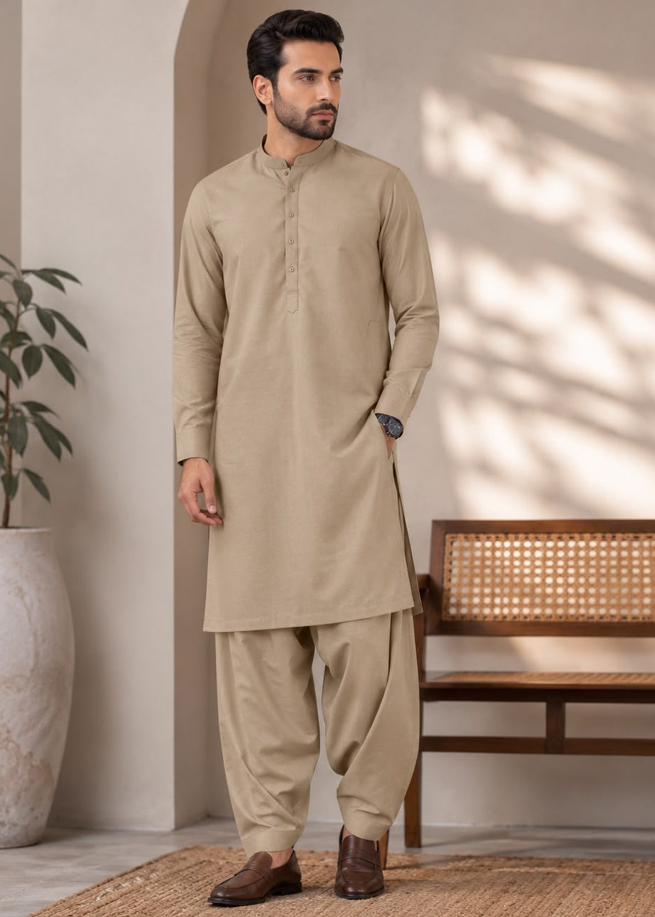 A man modeling a beige long-sleeve kurta and matching pants set in a styled indoor setting with a bench, potted plant, and natural light shadows.